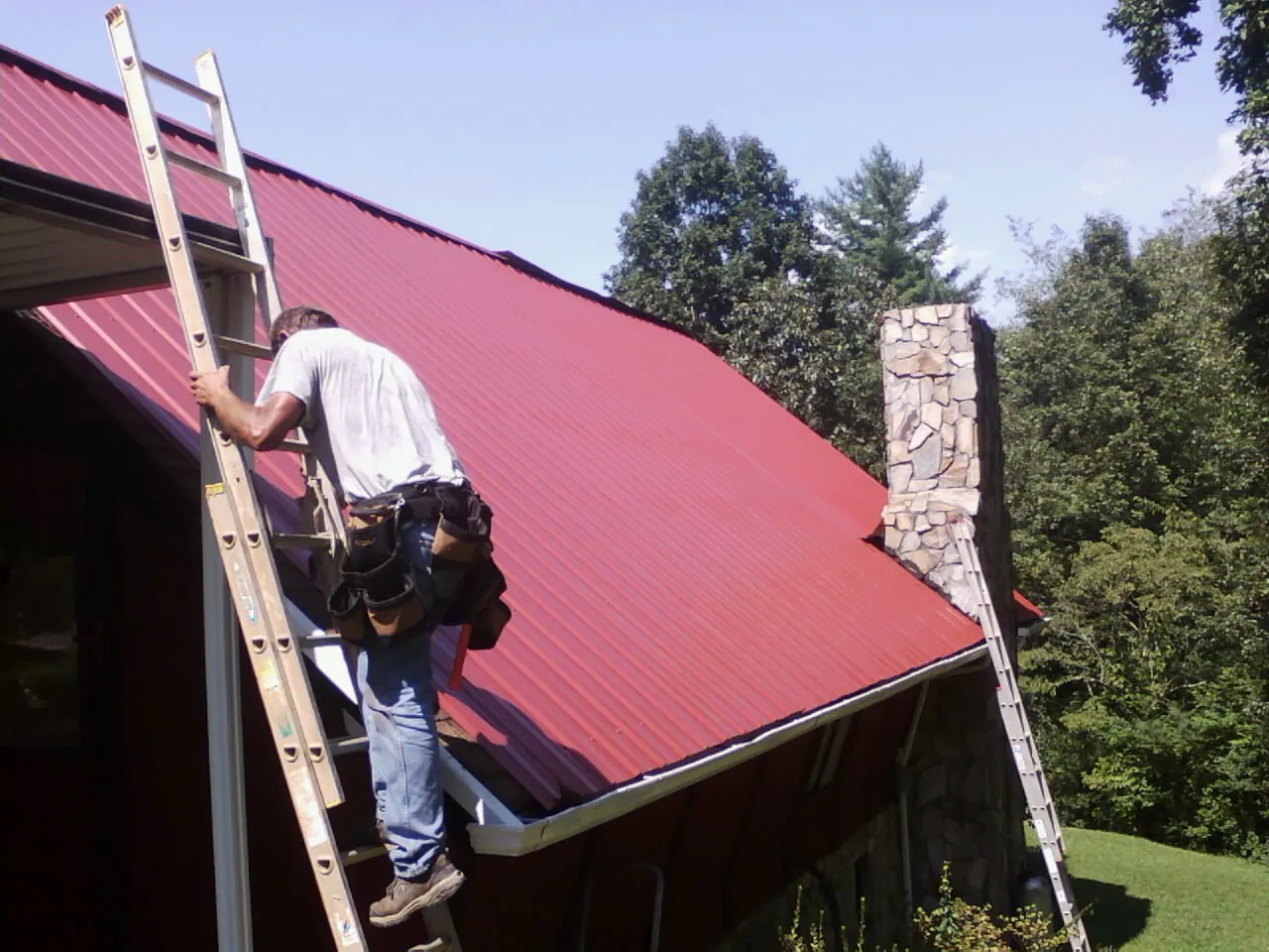Roofer on a ladder beside a red metal roof and stone chimney — Soffit & Fascia Repair by Roofing Brothers in Rutherford College